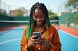 © miss irine - Young woman with braided hair smiles while using phone on tennis court. Casual style girl texting outdoor. She enjoys connection with people online.