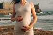 © SValeriia - Woman holds her baby during sunset with waves in background and warm sunlight shining.  mother stands on rocky beach at sunset. girl holds her pregnant belly . sun reflects off water, enjoys moment.