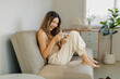 © amnaj - Young Asian woman smiling, laughing, and enjoying mobile gaming or social media app on phone while relaxing on a couch