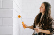 © Westend61 - Woman decorating a white brick wall indoors with a paint roller