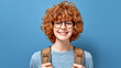 © Chainat - Smiling Teenager with Backpack A cheerful teenager with curly red hair, glasses, and a backpack radiates positivity against a vibrant blue backdrop. Perfect for representing youthful energy.