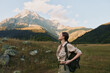 © SHOTPRIME STUDIO - Young woman traveler with backpack enjoys mountain landscape during sunny day, standing in grassy field with rocky peaks and blue sky in background, lifestyle concept.