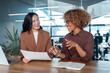 © Stock 4 You - Two businesswomen collaborating at modern office desk smiling during meeting with laptop and documents. Multiracial women talking working together. Teamwork and professional communication at workplace