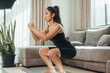 © sarayutsridee - Asian woman in black sportswear practicing bodyweight squats on a yoga mat in her living room. Side view of a fit woman doing a lower body workout at home. Concept of home fitness.