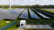 © AmazingAerialAgency - Aerial view of rows of solar panels in a large solar farm with a wind turbine and green fields under a cloudy sky in Warmenhuizen, North Holland, Netherlands.