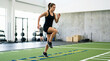 © Xenia - Female athlete performing footwork drills on an agility ladder. Woman in black activewear training on green turf in a modern gym