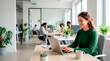 © Xenia - Smiling young adult businesswoman working on laptop in modern office. Happy female employee typing at desk with colleagues in background