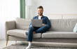 © Prostock-studio - Handsome bearded man using laptop and smiling, sitting on couch in living room, websurfing or using social media, copy space. Cheerful middle aged man enjoying part-time remote job