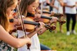 © Viktor - Close up of young girls playing violins outdoors at a summer festival. Children practice music lesson together, string instruments creating beautiful melodies in park.