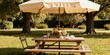 © Viktor - Wooden picnic table set for lunch under large umbrella in sunny park. Floral arrangement adorns table with cups and glasses. Chairs placed for outdoor dining.