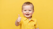 © julijadmi - Smiling toddler in yellow shirt giving thumbs up gesture against a bright yellow background, showcasing a cheerful expression and playful demeanor