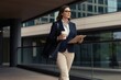 © Maryna - Professional woman walks near modern office building with coffee cup and clipboard. Confident lady in smart casual wear heading to work, busy city life concept.