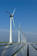 © Amazing Aerial - Aerial view of a row of tall wind turbines integrated with a large-scale solar panel array under a bright blue sky in the Netherlands.