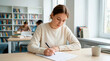 © Xenia - Focused female college student writing an essay at a desk in a library. Young woman concentrating on an exam paper in a university classroom. Higher education and academic success concept