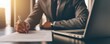 © Nart - A businessman in a suit writes on important paperwork beside a laptop in a sunlit office. The image highlights corporate planning and professional documentation.