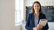 © dinda - Portrait of a confident smiling female teacher in a navy blazer holding books and notebooks in a modern classroom setting.