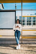 © BullRun - Young woman with dental braces holding smartphone and looking away, career growth, ambition, successful digital nomad waiting at city bus stop, wearing white sweater and jeans, urban lifestyle.