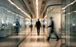 © Vhagar - A blurred image of professionals walking in a glass-walled office corridor, showcasing modern corporate architecture and a busy work environment.