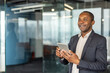 © Liubomir - Professional man confidently smiling, holding a digital tablet in a modern corporate office environment, actively engaging with technology and feeling successful