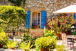 © Irina Schmidt - Sunny courtyard in Provence France with stone house, blue shutters and blooming flowers, cozy garden terrace, charming rural scene.