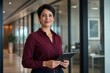© VeerSaif - Confident businesswoman in a modern office hallway, holding a digital tablet. Professional female executive, smiling and looking at the camera, representing leadership and success in corporate