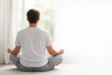 Man sitting in a lotus position on the floor, practicing meditation in a bright room with white curtains and natural light from a window
