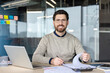 © Liubomir - Smiling bearded man with glasses at office desk, holding a pen while reviewing paperwork beside a laptop, confident professional planning and analyzing financial documents