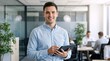 © Vasiliy - Smiling young businessman holding a digital tablet in a modern office. Professional male employee in a corporate workplace environment