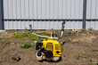 © WoodHunt - Yellow gasoline brush cutter engine unit on dry ground with weeds against a grey metal fence