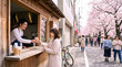 © Luka - Smiling male barista serving ice coffee to a woman at a wooden takeaway cafe window on a spring city street, blooming cherry blossom trees and people walking, small business and lifestyle concept