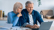 © Prostock-studio - Two older adults are seated at a table filled with papers, looking at a laptop. Their expressions are focused as they review financial documents together. Sunlight fills the room.