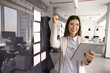 © fizkes - Happy young Hispanic woman holding tablet, receiving great news, celebrating sales growth or progress with clenched fist stands next to glass wall with attached sticky notes. Triumph, business success