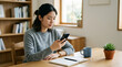 © NanisHappy - Woman using smartphone in quiet home office with notebook, mug, plant, and bookshelf