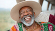 © Oksana - A joyous, senior man with a grey beard wearing a wide-brimmed hat and a vibrant, patterned shirt laughs heartily while seated inside a vehicle, with a blurred natural landscape visible through the win
