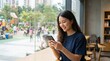 © kitti - Smiling young woman in a blue t shirt using her smartphone while sitting by a window in a cafe.