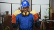 © Krakenimages.com - Man in blue hardhat and coveralls smiling with yellow ear defenders and heavy gloves points forward with gloved hands in building; confidence safety teamwork.
