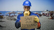 © Krakenimages.com - Man in blue hardhat and work gloves holds cardboard sign reading save our democracy and points finger toward camera on crowded beach; urgent plea.