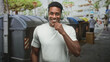 © Krakenimages.com - Man wearing headphones and white tshirt touches chin while smiling in front of large dumpsters on a busy street; joy.