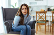 © Dragana Gordic - Young Woman Sitting at Home With Stomach Pain and Worried Expression in Living Room