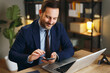 © djile - Happy businessman using a smartphone while working on a laptop at his office desk