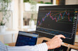 © Studio Marmellata - A person analyzes financial charts on multiple screens, pointing at a stock market graph on a large monitor.