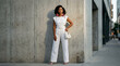 © Guillotine - Young Black woman posing in stylish white outfit against concrete wall, wearing leaf-shaped earrings