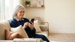 © spyrakot - Senior woman using smartphone on cozy living room couch
