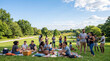 © MystoganCreative - A diverse group of friends enjoying a sunny outdoor picnic in a lush green park with trees and a pond.