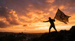 © solavie - Silhouette of a person holding a flag on a mountaintop against a dramatic sunset sky, representing triumph and achievement