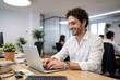 © Vasiliy - Smiling young man working on laptop in modern office. Professional male employee typing at desk. Business and technology concept