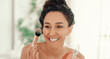 © Prostock-studio - A woman is applying makeup in a bright room. She smiles while holding a brush in one hand and a compact in the other. The setting has soft lighting with greenery in the background.