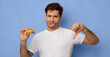 © Prostock-studio - A man stands with a burger in one hand and gestures a thumbs down with the other hand. He has a playful expression on his face. The background is a solid blue color.