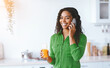 © Prostock-studio - A woman stands in a kitchen while talking on her phone. She holds a glass of orange juice in her hand. The kitchen has a bright and modern look. It is daytime.