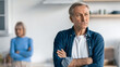 © Prostock-studio - A man stands with crossed arms, looking thoughtful in a kitchen. Behind him, a woman has her arms crossed and appears upset. The kitchen has modern features and a neutral color scheme.